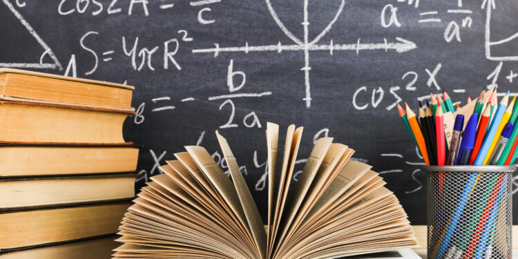 School desk in the classroom, with books on background of chalk board with written formulas. Soncept Teacher's Day.